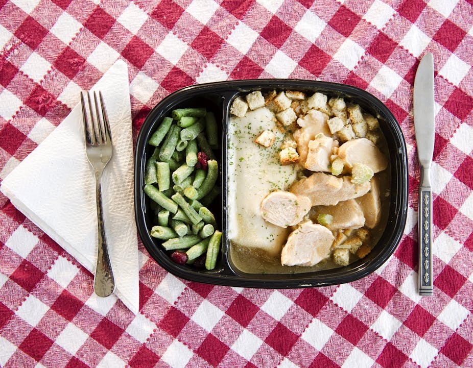 Close-up view of a TV dinner containing green beans, a white meat and other sides, sitting on a red checkered tablecloth.