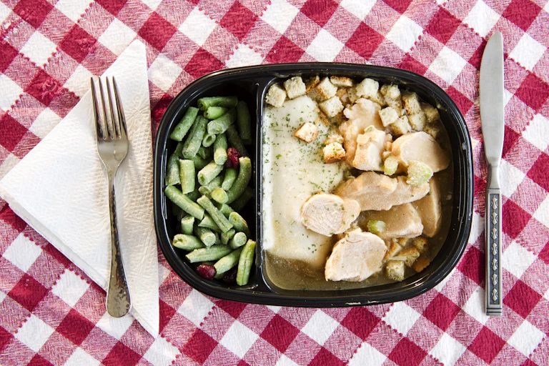 Close-up view of a TV dinner containing green beans, a white meat and other sides, sitting on a red checkered tablecloth.