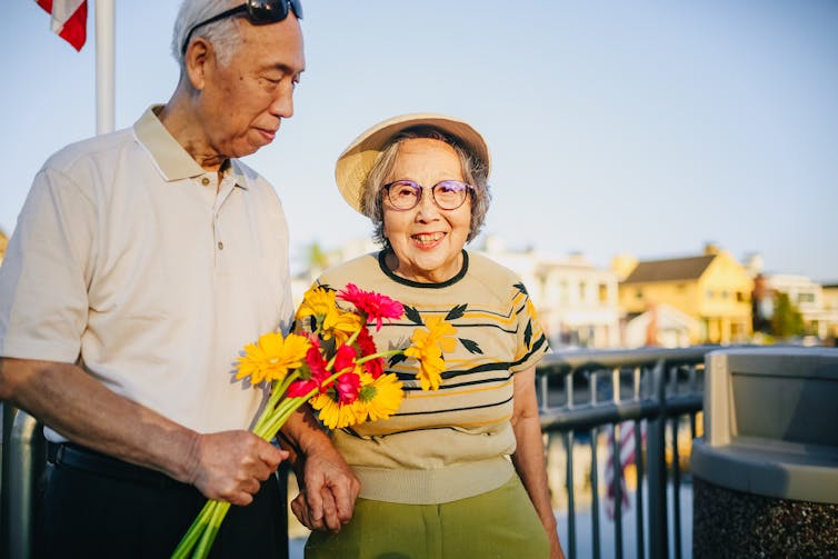 An older man looks lovingly at a smiling older woman. He holds a bunch of flowers towards her.