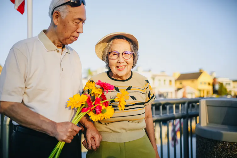 An older man looks lovingly at a smiling older woman. He holds a bunch of flowers towards her.