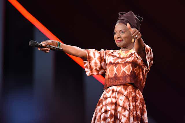A woman in a dress of African print and a head wrap smiles as she holds a microphone out to the audience with one hand and makes a thumbs up gesture with the other hand.
