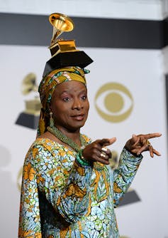 A woman in African attire points her fingers to the room as she poses for a photo with a gold statue of a gramophone balancing on her head.