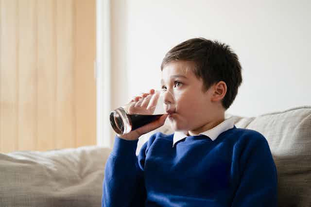 A boy sitting on the couch drinking a glass of soft drink.