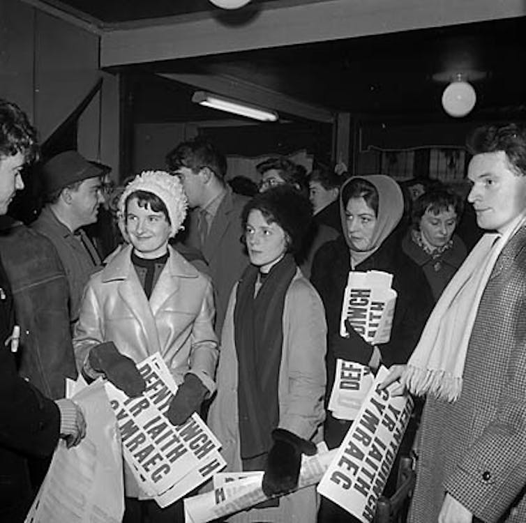 Black and white image of people in scarves, hats and coats carrying Welsh language signs saying 'defnyddiwch yr iaith Gymraeg'