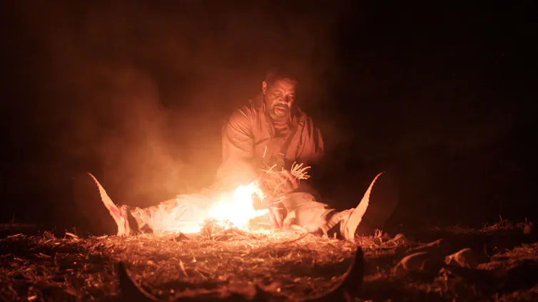 An elderly man sits at a fire, surrounded by darkness.