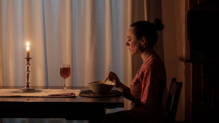 A woman sits eating dinner by candlelight, a glass of wine in front of her plate of food.