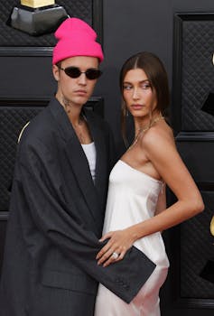 Justin Bieber wears a black suit and a pink beanie hat, holding the hip of his wife Hayley who wears her hair in a low ponytail and a strapless white dress.