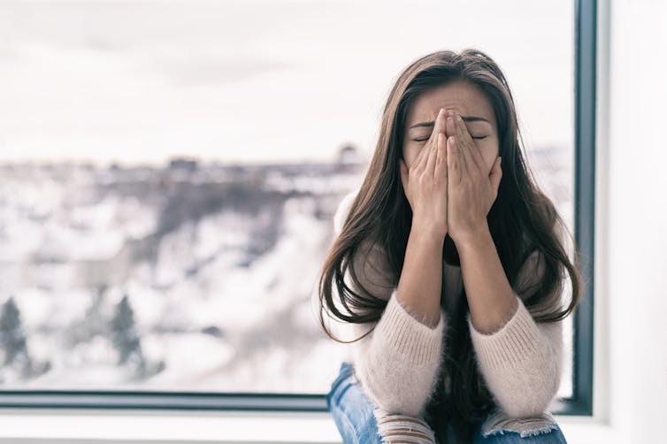 A woman sitting on a windowsill with her hands over her face.