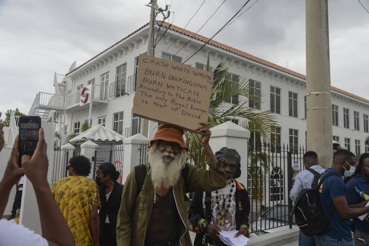 A Black man with a bushy grey beard holds up an anti-monarchy sign outside a white stone building.