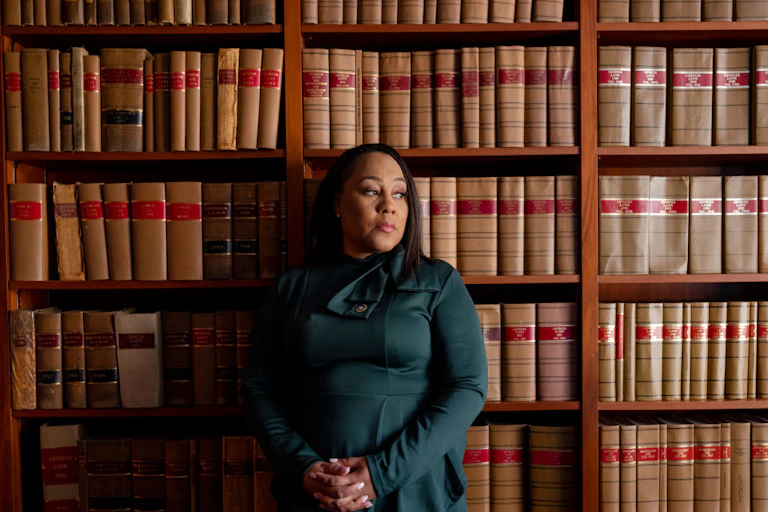A Black, middle aged woman in a dark green, formal outfit poses, looking to the side, in front of a tall bookshelf.