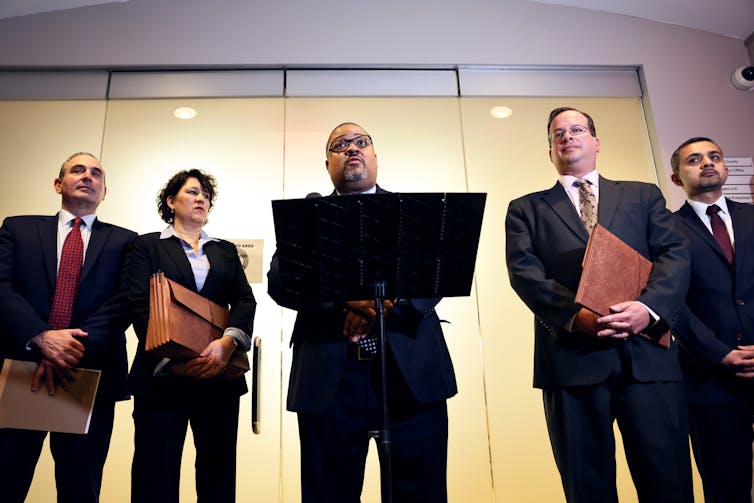 Five people, all in suits, stand in a row. One person in the center, a Black middle aged man, stands at a podium.