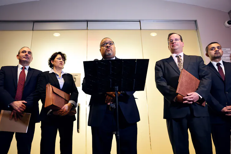 Five people, all in suits, stand in a row. One person in the center, a Black middle aged man, stands at a podium.