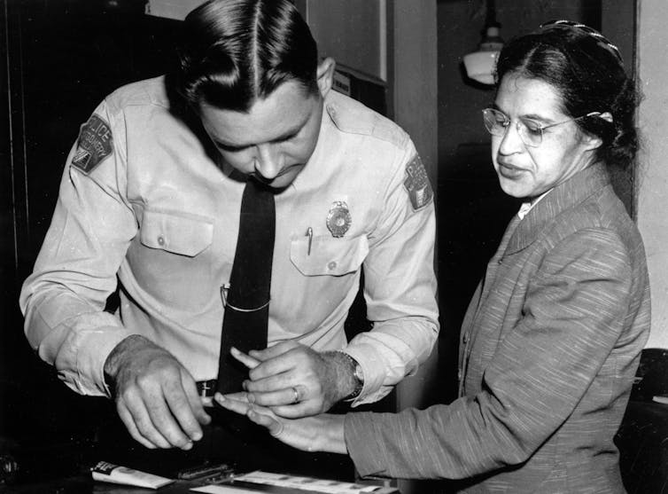 A Black woman is fingerprinted by a police officer in a black-and-white photo.