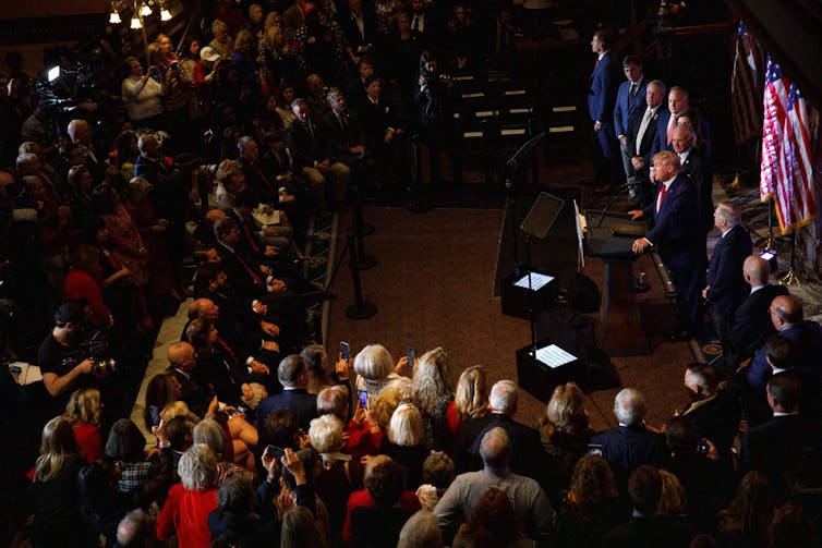 A large group of people are seen in an audience in a dark room, looking towards a white, older man in a suit speaking at a podium, with other men and American flags behind him.