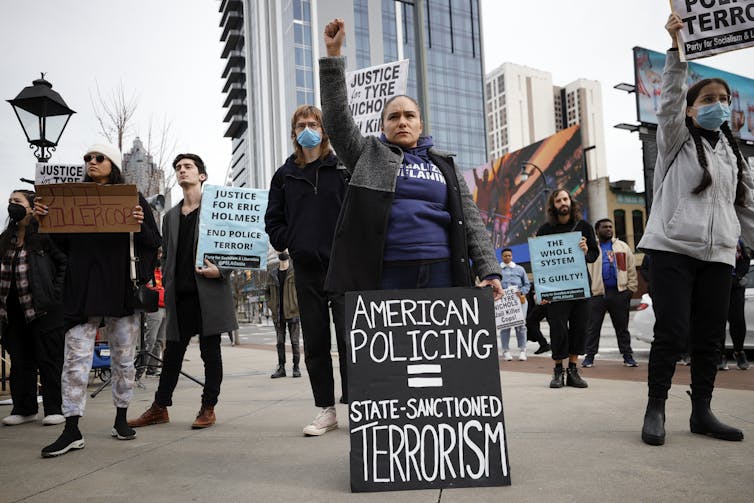 Protesters stand together, one with her arm raised holding a sign that says American Policing equals State-Sanctioned Terrorism.