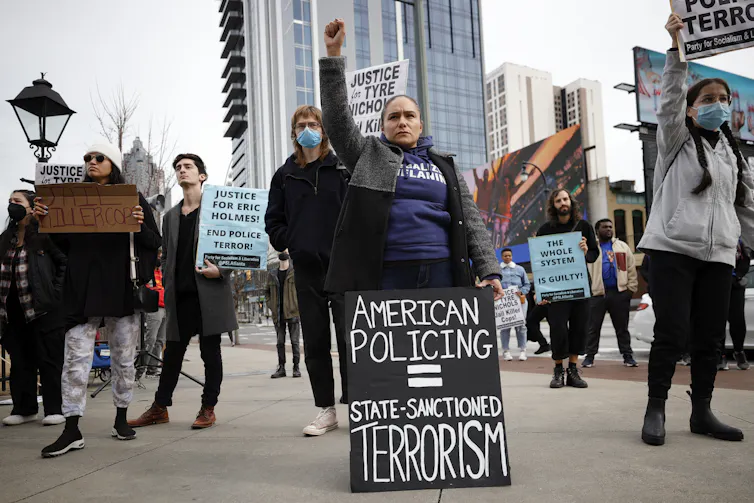 Protesters stand together, one with her arm raised holding a sign that says American Policing equals State-Sanctioned Terrorism.