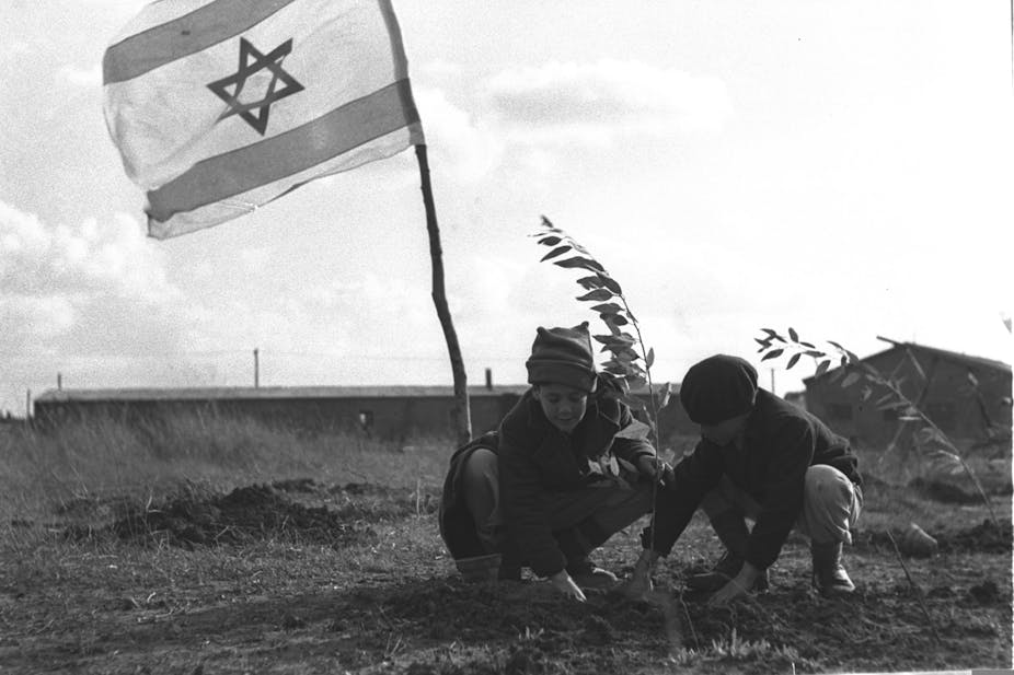 Two children in coats and caps plant a small tree in front of an Israeli flag.