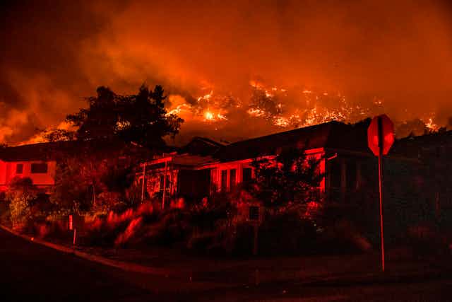 Wildfire burns in the hills above houses on a street with a stop sign.
