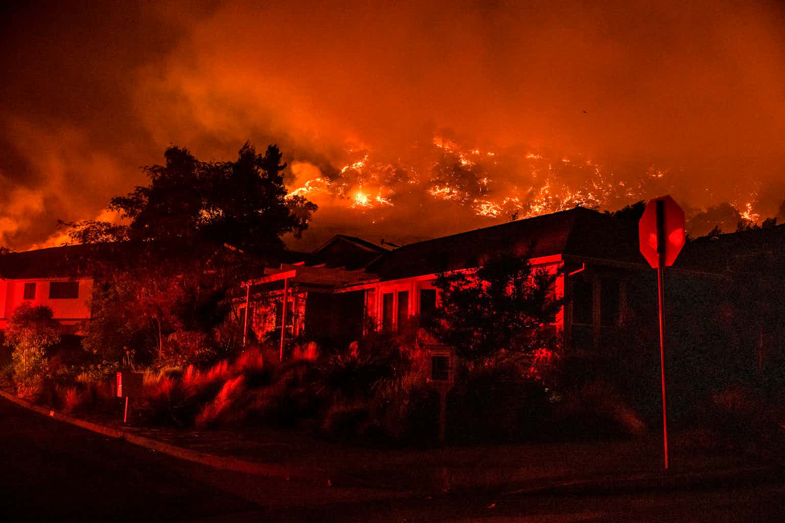 Wildfire burns in the hills above houses on a street with a stop sign.