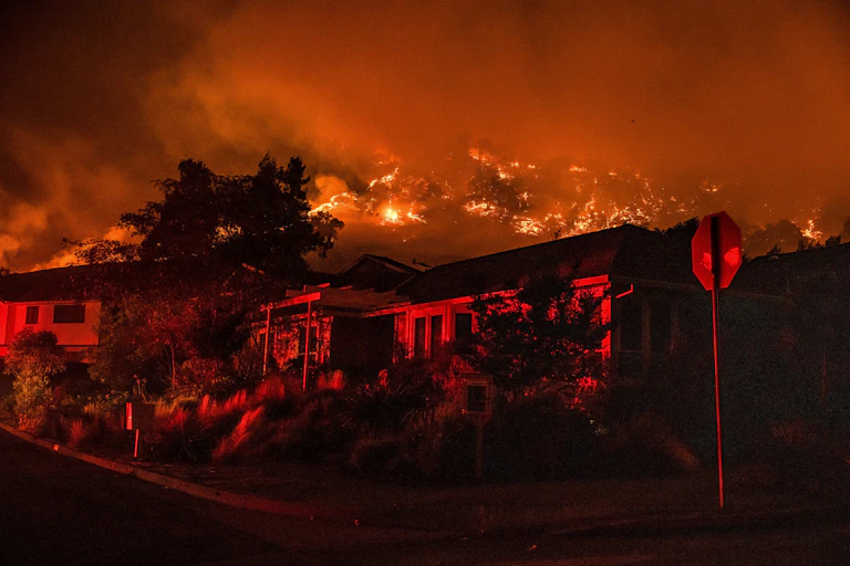 Wildfire burns in the hills above houses on a street with a stop sign.