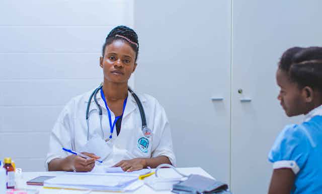 A Black woman in a white doctor's robe sits at a table and stares at the camera, a stethoscope around her neck and a pen in her hand.