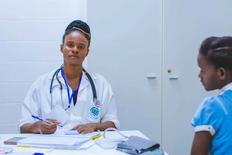 A Black woman in a white doctor's robe sits at a table and stares at the camera, a stethoscope around her neck and a pen in her hand.