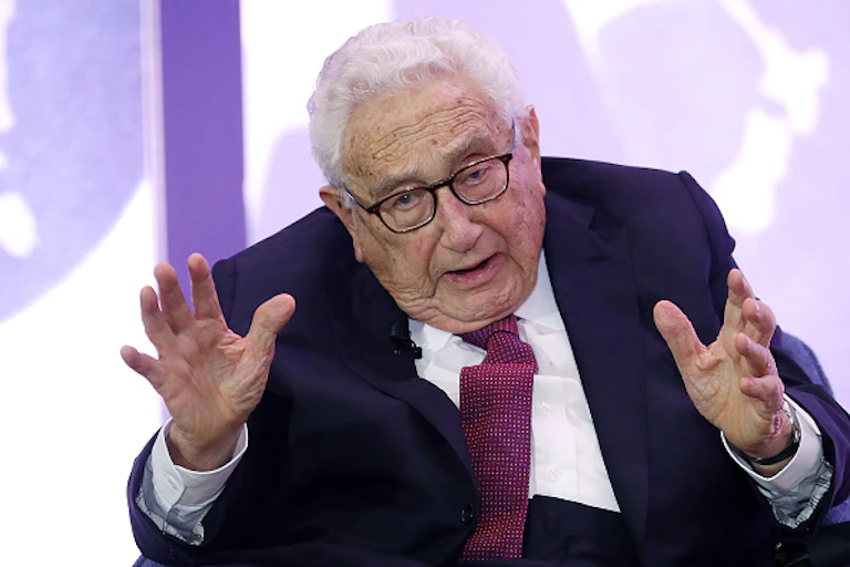 An old man wearing a jacket, tie and specs gesticulates with his arms, with his back hunched, while giving a speech.