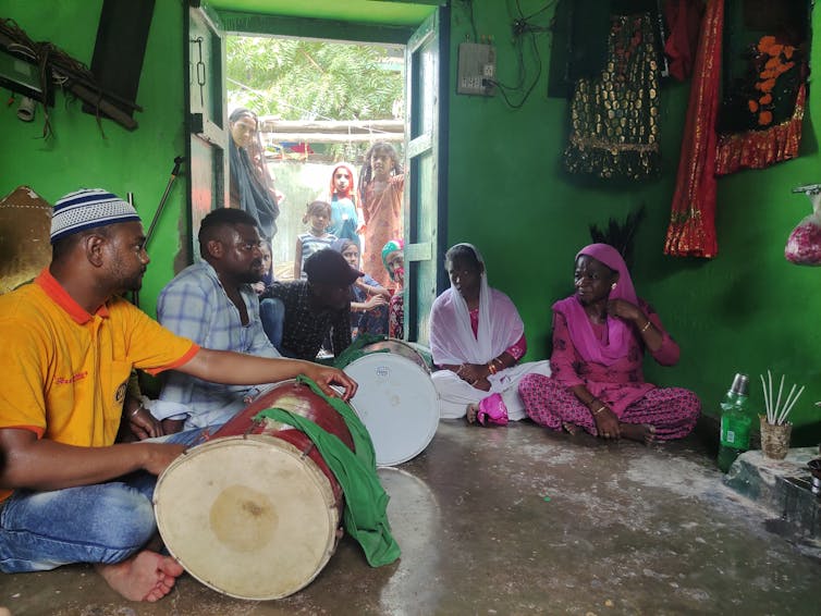 A group of people sit in a circle, some drumming on large drums. The doorway to the room is crowded with young observers.