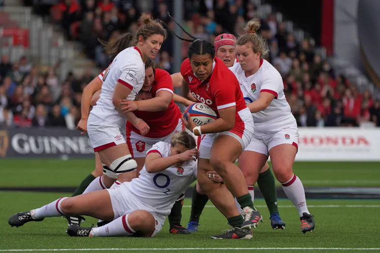 A female rugby player in red charges through a pack of rugby players in white with the ball under her arm.