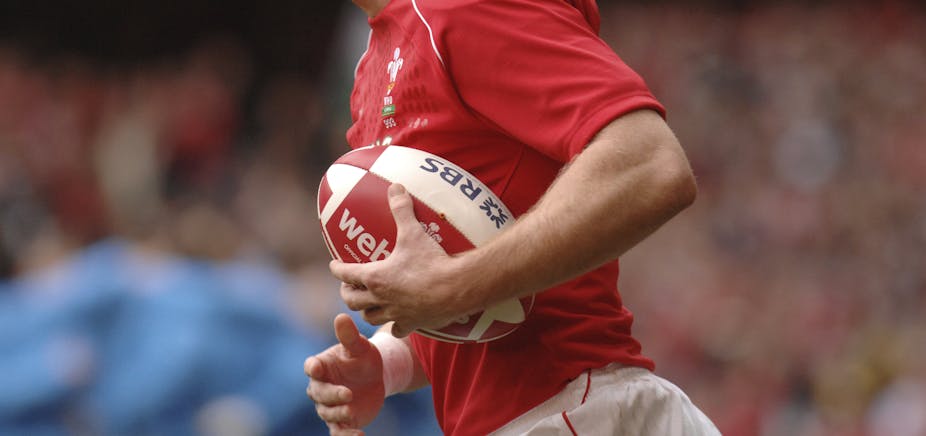 A close up of a rugby ball, held under the arm of a player wearing a red Wales shirt. 