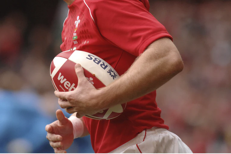 A close up of a rugby ball, held under the arm of a player wearing a red Wales shirt.