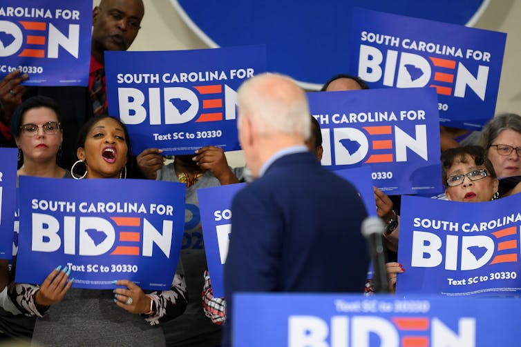 A suited man stands with his back to the camera, facing an ethnically diverse group of men and women holding signs that read 'South Carolina for Biden during the 2020 presidential primary process.'