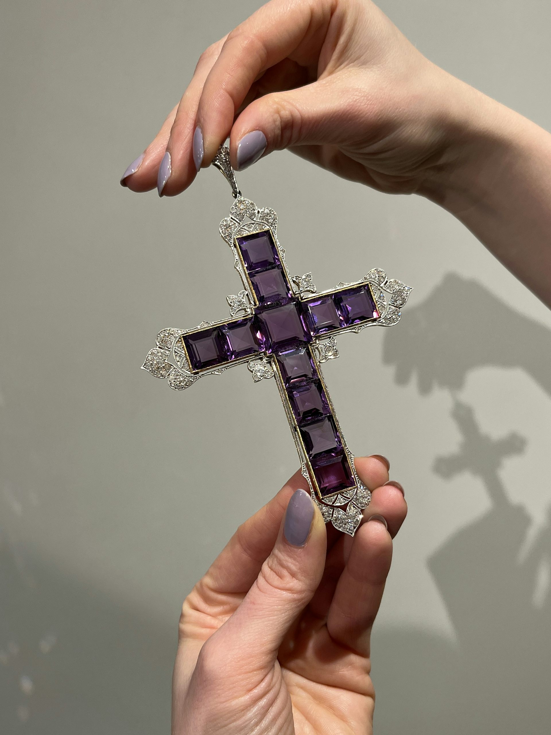 Hands holding the Attallah cross, constructed of purple square gems and encrusted with diamonds around the edges.