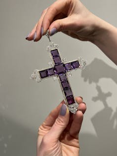 Hands holding the Attallah cross, constructed of purple square gems and encrusted with diamonds around the edges.