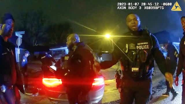 Police officers in uniform are captured on an video image in front of a car.