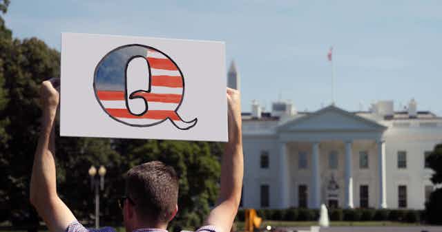 A man with a big Q sign stands outside the White House.