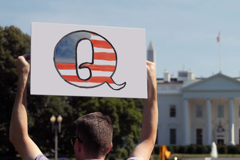 A man with a big Q sign stands outside the White House.