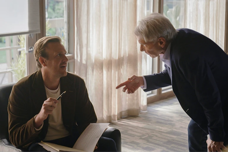 A man sits with a notepad and pen and speaks to a man who is stading.