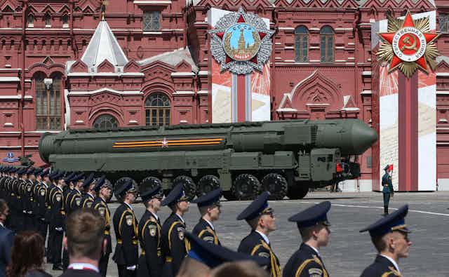 A row of soldiers in a dark uniform and hats face towards a large green missile, rolling in front of a red building. 