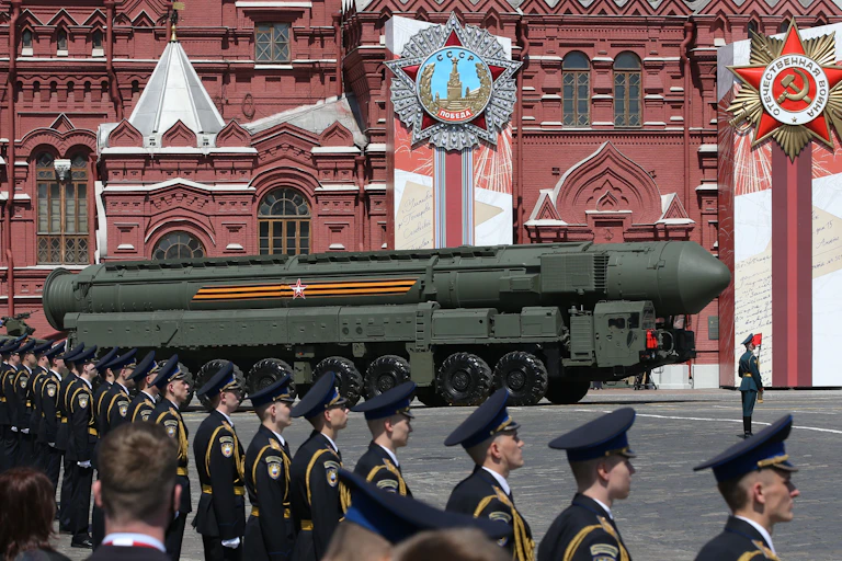 A row of soldiers in a dark uniform and hats face towards a large green missile, rolling in front of a red building.