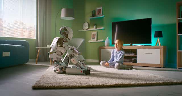 a humanoid robot and a girl face each other while sitting on rug in a living room
