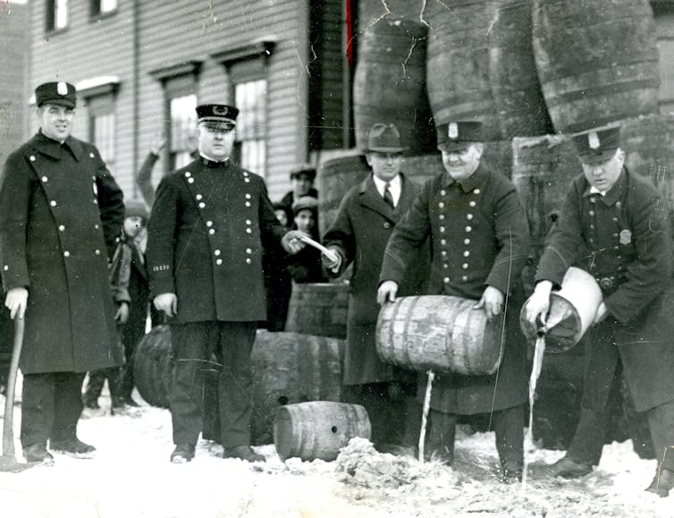 Black and white photo shows police officers in 1920s uniforms pouring out liquid from a barrel.