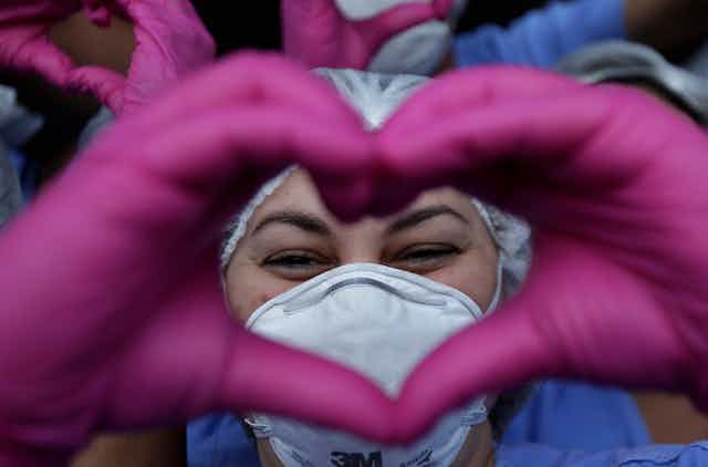 Close-up photo of a health worker in PPE making a heart sign with her hands in pink gloves