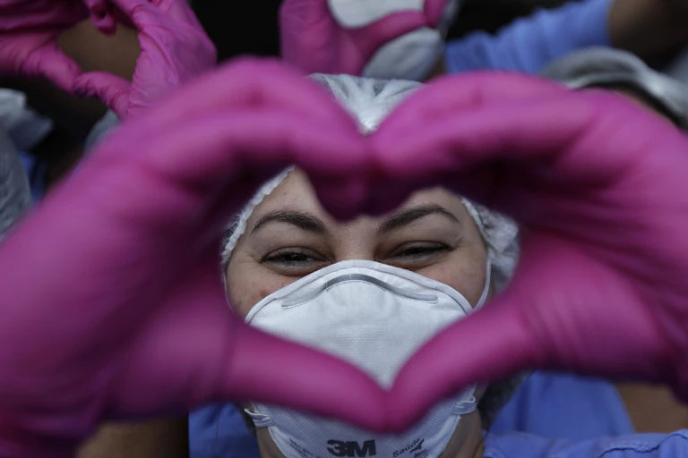 Close-up photo of a health worker in PPE making a heart sign with her hands in pink gloves