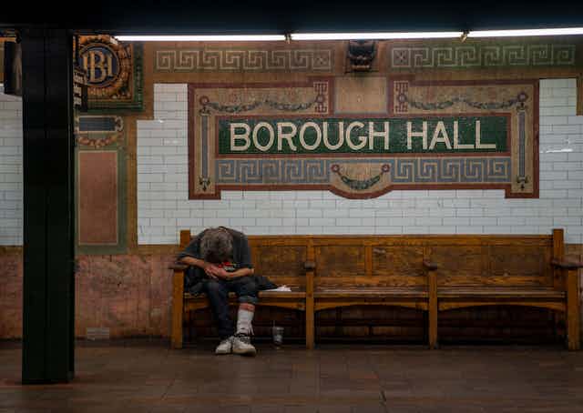 A person sits slumped over on an old bench inside a subway beneath a sign that says 'Borough Hall'