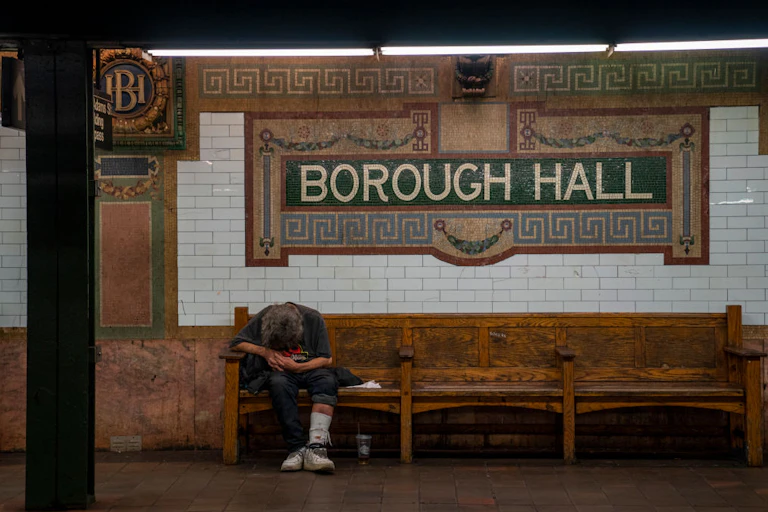 A person sits slumped over on an old bench inside a subway beneath a sign that says 'Borough Hall'