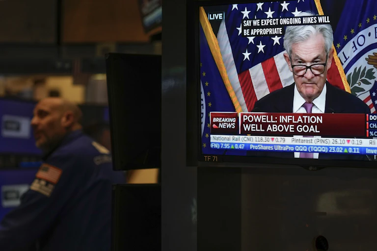 a white man with gray hair and glasses at the edge of his nose appears on a tv monitor with text as a man wearing a blue shirt looks on