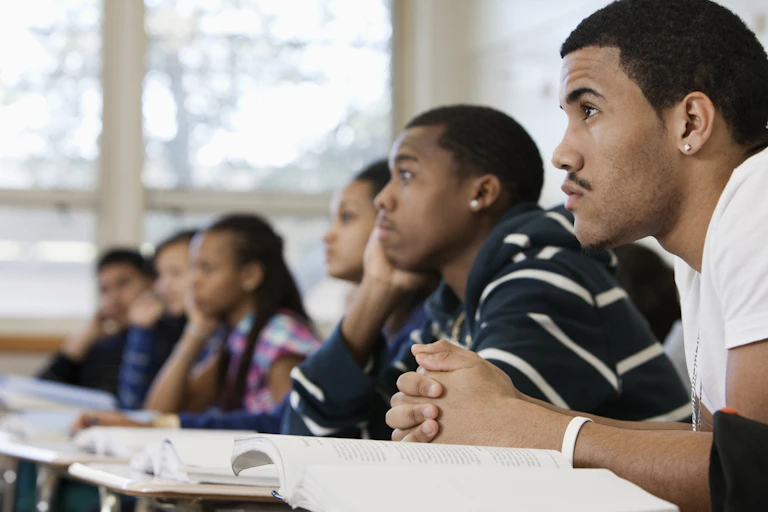A half dozen students from diverse backgrounds are shown from the side starting forward while seated in their desks.