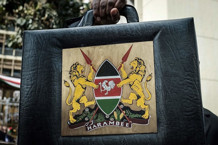 A black briefcase with a coat of arms in the middle showing two lions holding a shield with two spears criss-crossing it on the inside. The words 'Harambee' are written under the shield