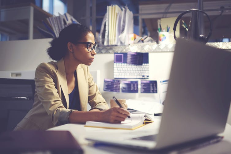 A young woman sits at a desk in a newsroom, taking notes and looking intently at a computer.